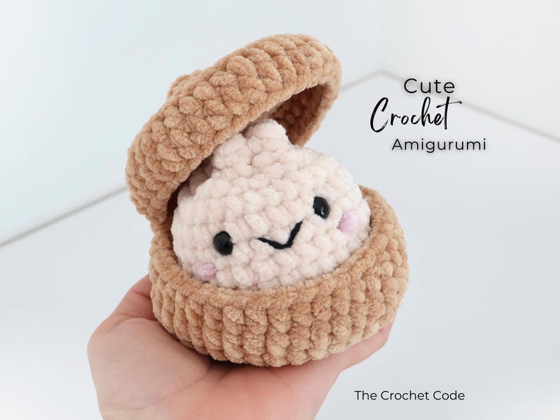 May include: A beige and cream crochet amigurumi dumpling inside a matching beige crochet container. The dumpling has black bead eyes, a stitched black mouth, and pink cheeks. Text reads: Cute Crochet Amigurumi and The Crochet Code.