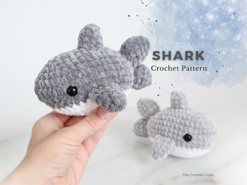 May include: Two gray crocheted shark toys with black eyes and white bellies. The text "SHARK Crochet Pattern" is displayed on the right side of the image. The smaller shark is on the right side of the image.