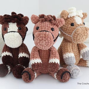 Three handmade crochet horse plushies in shades of brown, pink, and tan. Each plush horse has a white face and legs, with black accents for the eyes and bridle. The plushies are soft and cuddly, perfect for children.