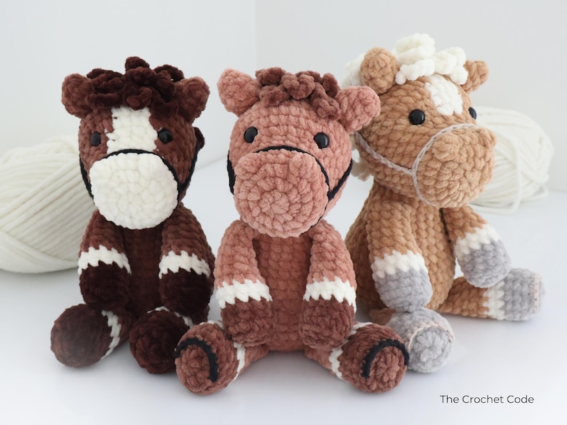 Three handmade crochet horse plushies in shades of brown, pink, and tan. Each plush horse has a white face and legs, with black accents for the eyes and bridle. The plushies are soft and cuddly, perfect for children.