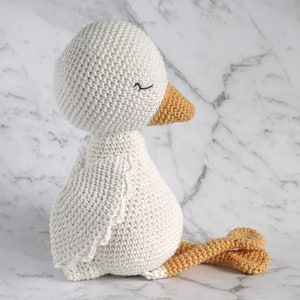 May include: A white crocheted goose with a yellow beak and feet. The goose is sitting on a white marble surface.