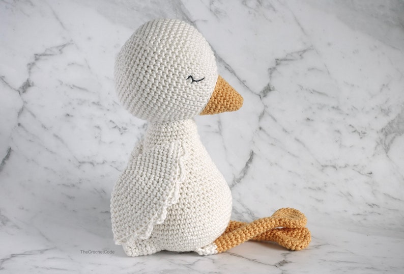 May include: A white crocheted goose with a yellow beak and feet. The goose is sitting on a white marble surface.