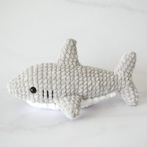 May include: A gray crocheted shark plush toy with white detailing around the belly. The shark has black eyes and a stitched mouth.