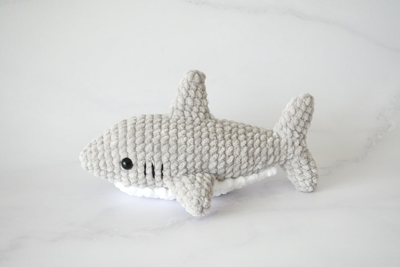 May include: A gray crocheted shark plush toy with white detailing around the belly. The shark has black eyes and a stitched mouth.