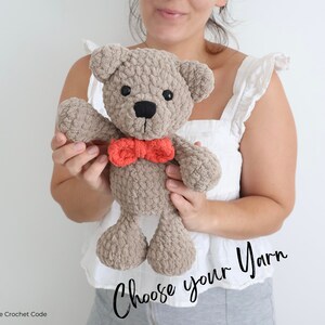 May include: A crocheted teddy bear with a brown body and a red bow tie. The bear is being held by a person. The text "Choose your yarn" is written in black cursive on the bottom of the image.