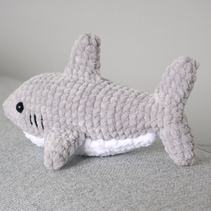 May include: A gray crocheted shark plush toy with a white belly and black eyes. The toy is lying on a gray surface.