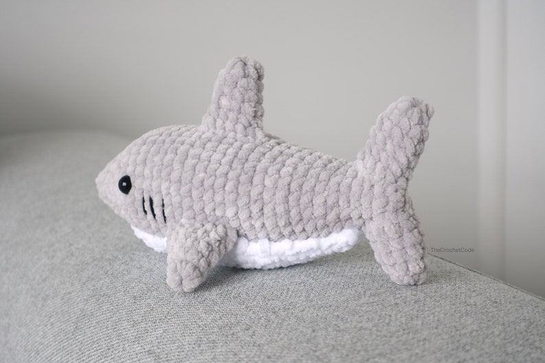 May include: A gray crocheted shark plush toy with a white belly and black eyes. The toy is lying on a gray surface.