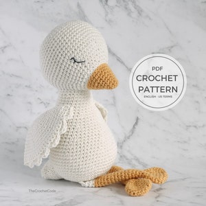 May include: A white crocheted goose amigurumi toy with a yellow beak and feet. The toy is sitting on a white marble surface. The text "PDF CROCHET PATTERN ENGLISH - US TERMS" is on a white circle.