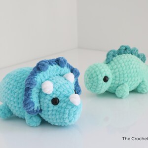 May include: Two handmade crochet dinosaur toys. One is a light blue triceratops with dark blue accents and white horns. The other is a small, mint green dinosaur. Both have black eyes and are made of soft yarn.