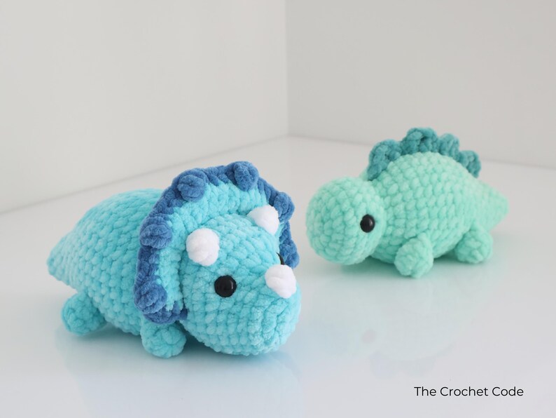 May include: Two handmade crochet dinosaur toys. One is a light blue triceratops with dark blue accents and white horns. The other is a small, mint green dinosaur. Both have black eyes and are made of soft yarn.