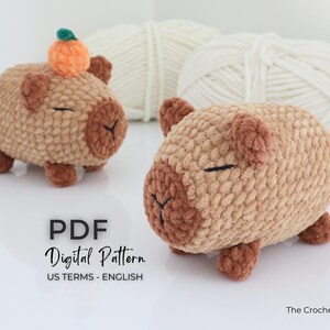 May include: Two crocheted capybara plushies in shades of brown, with small ears and closed eyes. One has an orange pumpkin on its head. The image includes text that reads "PDF Digital Pattern US TERMS - ENGLISH" and "The Crochet Code".