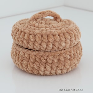 May include: A round, light brown crochet container with a matching lid and a small handle on top. The item is made from thick, soft yarn with a visible looped texture. The text "The Crochet Code" is printed in the bottom right corner of the image.