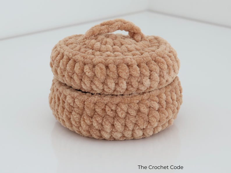 May include: A round, light brown crochet container with a matching lid and a small handle on top. The item is made from thick, soft yarn with a visible looped texture. The text "The Crochet Code" is printed in the bottom right corner of the image.