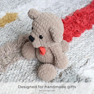 May include: A crocheted teddy bear with a red bow, sitting on a white and gray rug with a red and yellow stripe. The bear is made of a soft, fuzzy yarn and has a sweet expression.