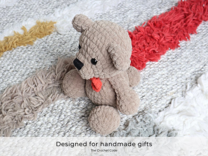 May include: A crocheted teddy bear with a red bow, sitting on a white and gray rug with a red and yellow stripe. The bear is made of a soft, fuzzy yarn and has a sweet expression.