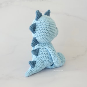 May include: A light blue crocheted dinosaur toy with blue accents. The dinosaur is sitting on a white surface.