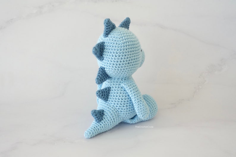 May include: A light blue crocheted dinosaur toy with blue accents. The dinosaur is sitting on a white surface.