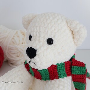 May include: A handmade, white crochet polar bear with black eyes and nose, wearing a red and green striped scarf. The bear is soft and plush, with a textured surface. The Crochet Code is printed on the bottom left.