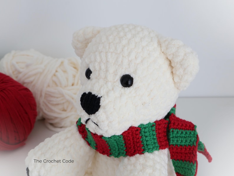 May include: A handmade, white crochet polar bear with black eyes and nose, wearing a red and green striped scarf. The bear is soft and plush, with a textured surface. The Crochet Code is printed on the bottom left.