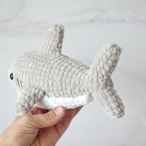 May include: A gray crocheted shark plush toy with white detailing on the belly. The toy is held in a hand.