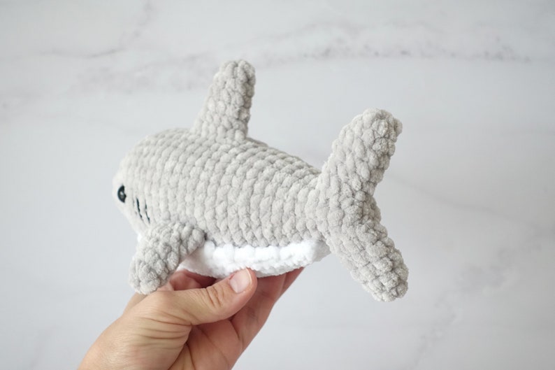 May include: A gray crocheted shark plush toy with white detailing on the belly. The toy is held in a hand.