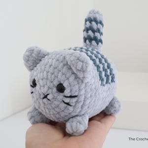 May include: A handmade, gray crochet cat with black eyes and whiskers. The cat has a striped pattern on its back and tail in a darker shade of gray. The Crochet Code is printed on the bottom right.