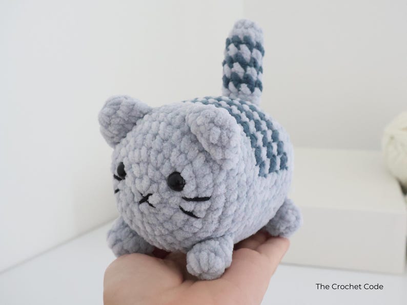 May include: A handmade, gray crochet cat with black eyes and whiskers. The cat has a striped pattern on its back and tail in a darker shade of gray. The Crochet Code is printed on the bottom right.