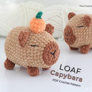 Könnte beinhalten: Zwei gehäkelte Capybaras, eines mit einem orangefarbenen Kürbis auf dem Kopf. Die Capybaras sind hellbraun mit braunen Akzenten und geschlossenen Augen. Der Text "LOAF Capybara PDF Crochet Pattern" ist sichtbar.