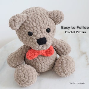 May include: A crocheted teddy bear with brown fur, black eyes, and a black nose. The bear is wearing a red bow tie and is sitting on a white surface. Easy to Follow Crochet Pattern.
