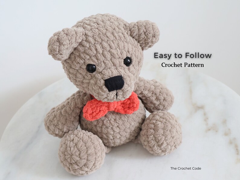 May include: A crocheted teddy bear with brown fur, black eyes, and a black nose. The bear is wearing a red bow tie and is sitting on a white surface. Easy to Follow Crochet Pattern.