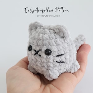 May include: A small, gray crocheted cat with black eyes and whiskers is held in a hand. The cat has a rounded body and small ears. The text "Easy-to-follow Pattern by TheCrochetCode" is at the top of the image.