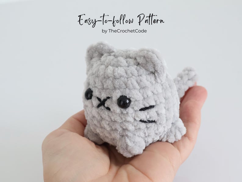 May include: A small, gray crocheted cat with black eyes and whiskers is held in a hand. The cat has a rounded body and small ears. The text "Easy-to-follow Pattern by TheCrochetCode" is at the top of the image.