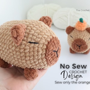 May include: Two crocheted capybara plushies in shades of brown and tan. One is larger and held in a hand, the other is smaller and has a pumpkin on its head. The text "No Sew Crochet Design" and "Sew only the orange!" is visible.