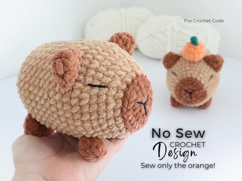 May include: Two crocheted capybara plushies in shades of brown and tan. One is larger and held in a hand, the other is smaller and has a pumpkin on its head. The text "No Sew Crochet Design" and "Sew only the orange!" is visible.