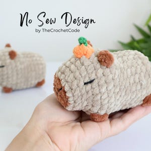 K&ouml;nnte beinhalten: Ein geh&auml;keltes Pl&uuml;schtier in Form eines Capybaras mit einem kleinen orangefarbenen K&uuml;rbis auf dem Kopf. Das Spielzeug ist braun und beige und hat eine weiche, flauschige Textur. No Sew Design von TheCrochetCode.