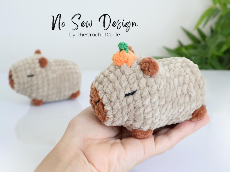 K&ouml;nnte beinhalten: Ein geh&auml;keltes Pl&uuml;schtier in Form eines Capybaras mit einem kleinen orangefarbenen K&uuml;rbis auf dem Kopf. Das Spielzeug ist braun und beige und hat eine weiche, flauschige Textur. No Sew Design von TheCrochetCode.