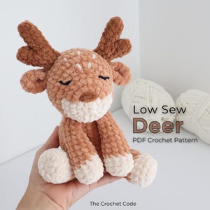 May include: A crocheted deer toy in brown and cream. The deer has a soft, textured appearance with a white muzzle and belly, and black eyes. The image includes text that reads "Low Sew Deer PDF Crochet Pattern".
