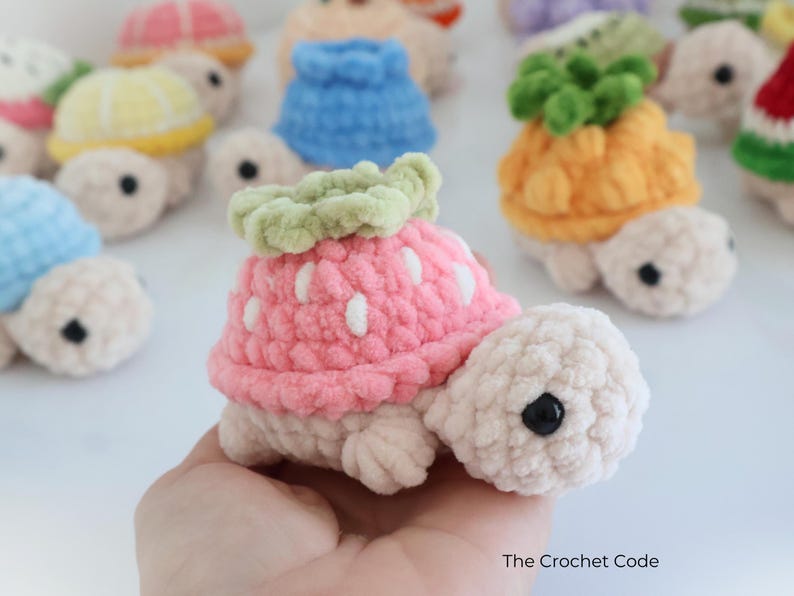 May include: A collection of handmade crochet turtles, each with a unique fruit-themed shell. The featured turtle has a pink strawberry shell with white accents and a green leafy top. Other turtles have shells resembling a pineapple, lemon, and watermelon.