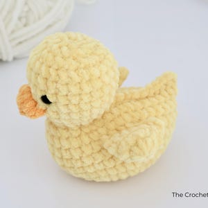 May include: A handmade, yellow crochet duck with an orange beak and black eyes. The duck is made of soft, textured yarn and is sitting on a white surface. A ball of white yarn is in the background. The Crochet Code is written on the bottom right.