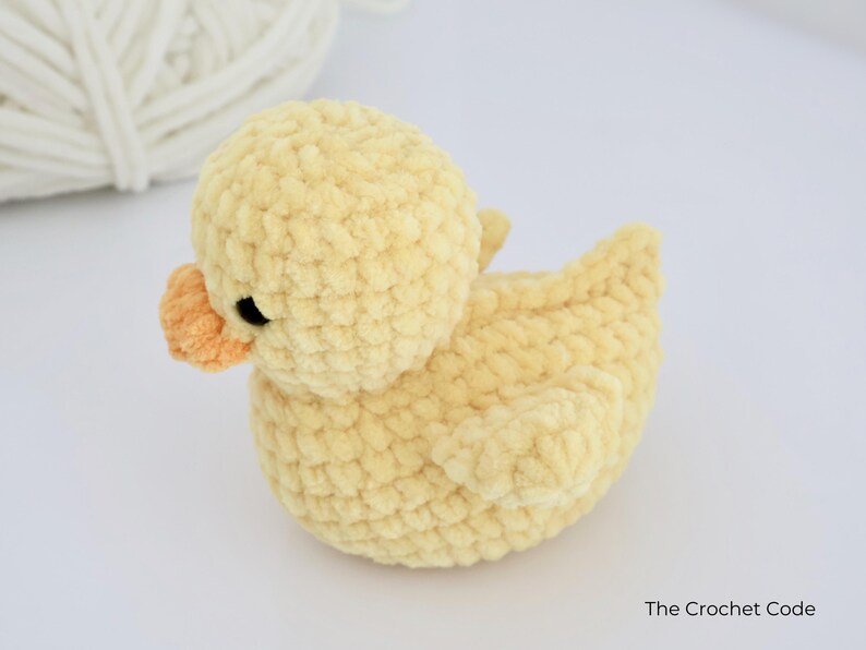 May include: A handmade, yellow crochet duck with an orange beak and black eyes. The duck is made of soft, textured yarn and is sitting on a white surface. A ball of white yarn is in the background. The Crochet Code is written on the bottom right.