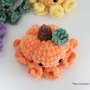 Puede incluir: Un peluche de pulpo con forma de calabaza naranja hecho de hilo suave con un tallo marr&oacute;n y una hoja verde en la parte superior. Tiene ojos de pl&aacute;stico negro y tent&aacute;culos rizados. Se ven otros peluches de pulpo similares en morado, verde y amarillo.