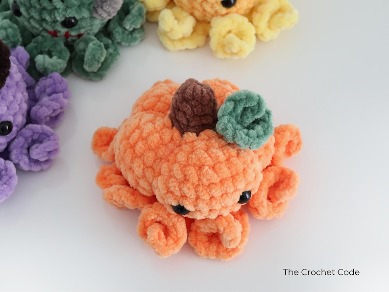 Puede incluir: Un peluche de pulpo con forma de calabaza naranja hecho de hilo suave con un tallo marr&oacute;n y una hoja verde en la parte superior. Tiene ojos de pl&aacute;stico negro y tent&aacute;culos rizados. Se ven otros peluches de pulpo similares en morado, verde y amarillo.