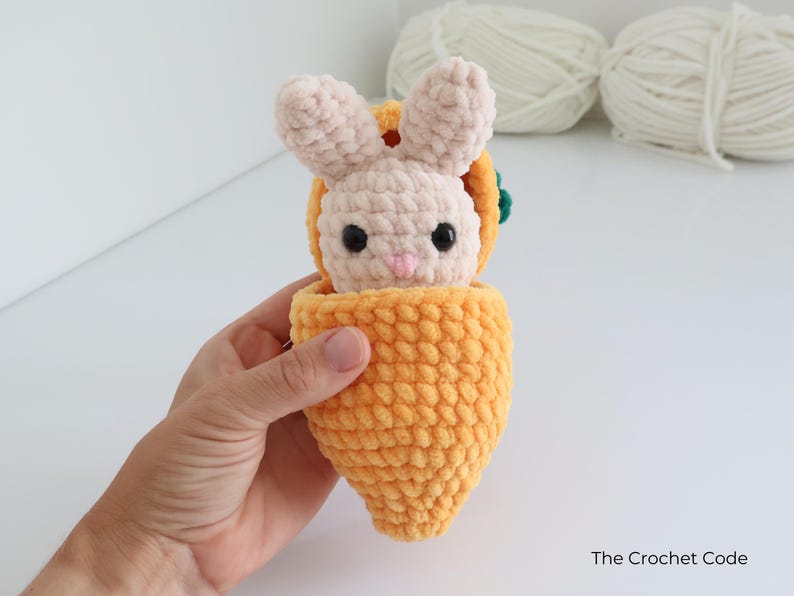 May include: A handmade crochet bunny toy in a carrot-shaped carrier. The bunny is light pink with black eyes and a pink nose. The carrot is bright orange with a green top. The Crochet Code is written on the bottom right.