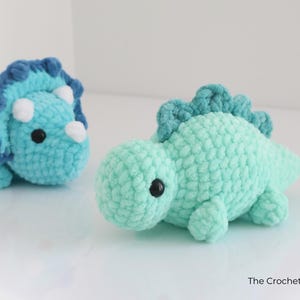 May include: Two crocheted dinosaur toys. One is a triceratops in light blue with dark blue accents and white horns. The other is a green dinosaur with a spiky back. Both have black safety eyes and are handmade.