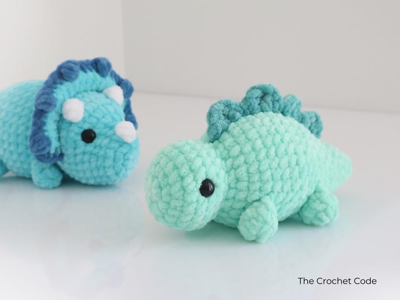 May include: Two crocheted dinosaur toys. One is a triceratops in light blue with dark blue accents and white horns. The other is a green dinosaur with a spiky back. Both have black safety eyes and are handmade.