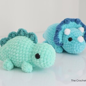 May include: Two handmade crochet dinosaur toys. One is a light teal color with a darker teal spine. The other is a triceratops in shades of blue with white horns. Both have black safety eyes. The Crochet Code is in the bottom right.