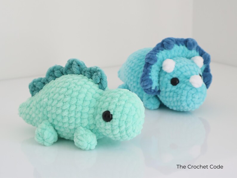 May include: Two handmade crochet dinosaur toys. One is a light teal color with a darker teal spine. The other is a triceratops in shades of blue with white horns. Both have black safety eyes. The Crochet Code is in the bottom right.