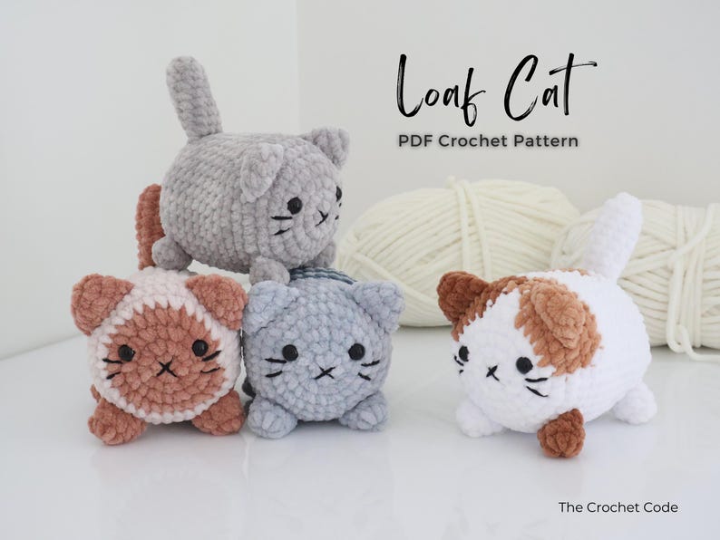May include: Four crocheted cat toys in various colors: gray, white with brown patches, and pink with brown patches. Each cat has black embroidered facial features. The text "Loaf Cat PDF Crochet Pattern" is displayed in the image.
