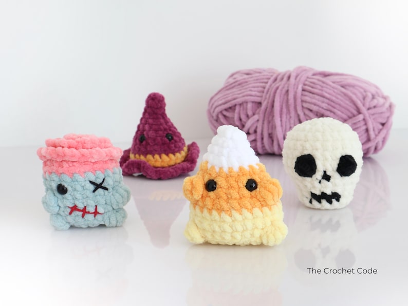 May include: A selection of handmade crochet Halloween figures. Includes a blue and pink monster, a purple witch hat, a candy corn, and a white skull. A ball of lavender yarn is in the background. The Crochet Code is written on the bottom right.
