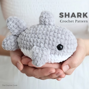 May include: A gray and white crocheted shark toy with black eyes and fins. The text "SHARK Crochet Pattern" is displayed above the shark. The Crochet Code is written at the bottom left.